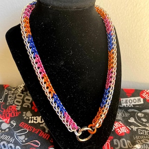 May include: A chainmail necklace constructed from interlocking metal rings in alternating colors of silver, bright pink, vibrant blue, and orange. The necklace features a circular metal ring clasp at the bottom, connecting the two ends of the chain.