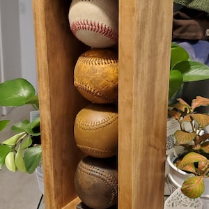 May include: A wooden display case holding four baseballs. The top baseball is white with red stitching, while the others are various shades of brown. The case is made of light brown wood.