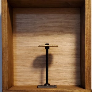 May include: A wooden display shelf with a light brown interior and a dark brown exterior. A metal and wood object is centered inside the shelf. The object has a black threaded rod, a small rectangular base, and a circular top.