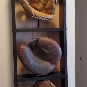Könnte beinhalten: Ein schwarzes Wandregal aus Holz mit drei Ebenen, auf denen jeweils ein alter Baseballhandschuh aus Leder steht. Das oberste Regal zeigt einen beigen Feldspielerhandschuh, das mittlere einen dunkelbraunen Catcher-Handschuh und das untere einen abgenutzten braunen Handschuh.