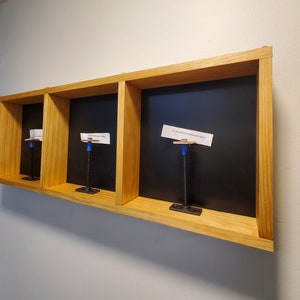 May include: A wooden wall shelf with three compartments, each with a black background and a white paper sign attached to a blue and black metal hook. The signs read "A little bit of whimsy goes a long way", "The future is now", and "A little bit of whimsy goes a long way".