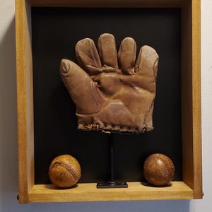 May include: A vintage baseball glove and two baseballs displayed in a wooden shadow box. The glove is brown and worn, mounted on a black background. The baseballs are also brown and show signs of wear.