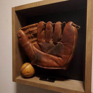 Peut inclure: Un gant de baseball et une balle vintage exposés dans une boîte d'ombre en bois. Le gant est en cuir marron, montrant des signes d'usure, et la balle est jaunie avec des coutures visibles. L'ensemble est monté sur un mur de couleur neutre.