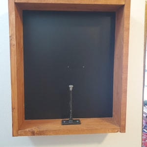 May include: A wooden shadow box with a dark interior and a metal support. The box is made of light brown wood and is mounted on a white wall. The metal support is attached to the bottom of the box.