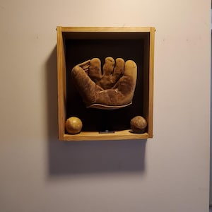 Può includere: Un guantone da baseball vintage in pelle marrone montato su un supporto nero all'interno di una cornice rettangolare in legno chiaro, accompagnato da due palline da baseball usurate posizionate sul bordo inferiore della teca.