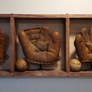 May include: Three vintage baseball gloves and two baseballs displayed in a wooden shadow box. The gloves are brown leather, and the baseballs are off-white with red stitching. The wooden frame has a dark finish.