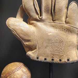 Peut inclure: Un gant et une balle de baseball anciens. Le gant est en cuir beige avec des coutures visibles et le texte "Genuine Cheeksksin" et "West Sporting Goods" est estampé sur le gant. La balle de baseball est également beige avec des coutures rouges.
