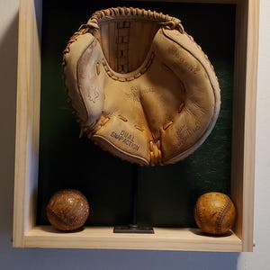 May include: A vintage baseball glove and two baseballs displayed in a wooden shadow box. The glove is tan leather with the text "Regent" and "Dual Snap Action". The baseballs are brown with red stitching.