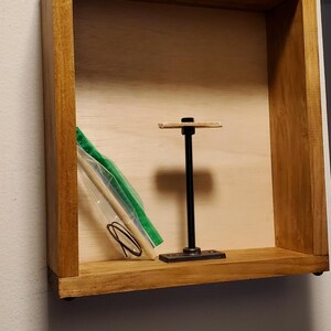 May include: Wooden square shelf with a light wood interior and a dark wood frame. Inside, a black metal display stand and a small bag with green and clear contents are visible. The shelf is mounted on a white wall.
