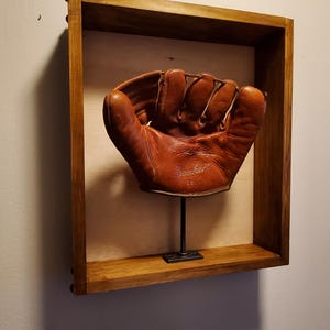 Peut inclure: Un gant de baseball en cuir marron exposé dans une boîte à ombre en bois avec un support en métal. Le gant porte l'inscription "Dembert".