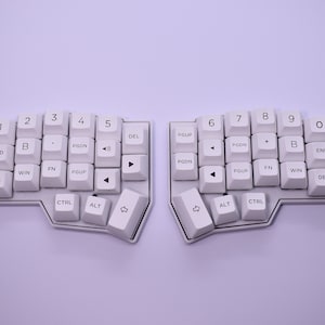 Può includere: Una tastiera bianca e nera divisa in due parti, con tasti etichettati con numeri, lettere e tasti funzione. La tastiera ha un design unico con una forma curva.