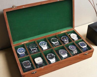May include: A wooden watch display case with a green felt interior, holding twelve assorted wristwatches. The case is open, showcasing the watches with various band colors and face designs. The case is made of a dark wood with brass hinges.
