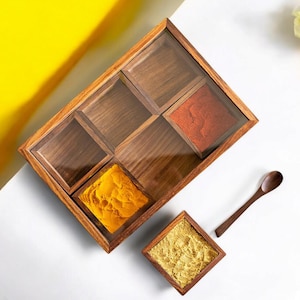 May include: A wooden spice box with a clear lid, containing various colourful spices. A small wooden spoon and a separate square spice container are also present. The spices include vibrant yellow and red powders. The box is set against a white and yellow background.