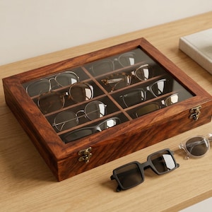 Könnte beinhalten: Eine Brillen-Vitrine aus Holz mit Glasdeckel, die verschiedene Brillenpaare zeigt. Das Gehäuse ist aus dunklem Holz gefertigt und hat messingfarbene Verschlüsse. Mehrere Sonnenbrillenpaare sind neben dem Gehäuse platziert.