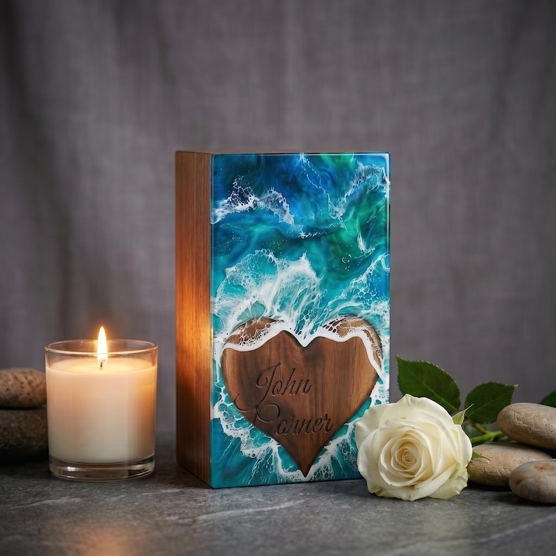 a wooden cremation urn with heart design on the front and filled with epoxy resin is kept on a grey countertop with flowers and candles.