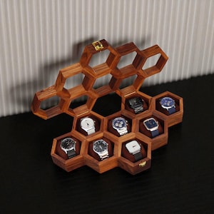 Puede incluir: Un expositor de relojes de madera con diseño de panal, que contiene varios relojes de pulsera. La caja es de madera oscura y tiene compartimentos individuales para cada reloj. Los relojes tienen varios estilos de correa y esfera.