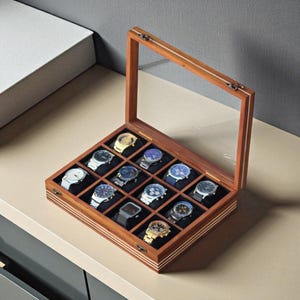 Puede incluir: Una caja de madera para exhibir relojes con tapa de cristal transparente, que muestra una colección de relojes de pulsera. La caja abierta revela doce relojes con diferentes diseños de correas y esferas, sobre un interior de terciopelo negro.