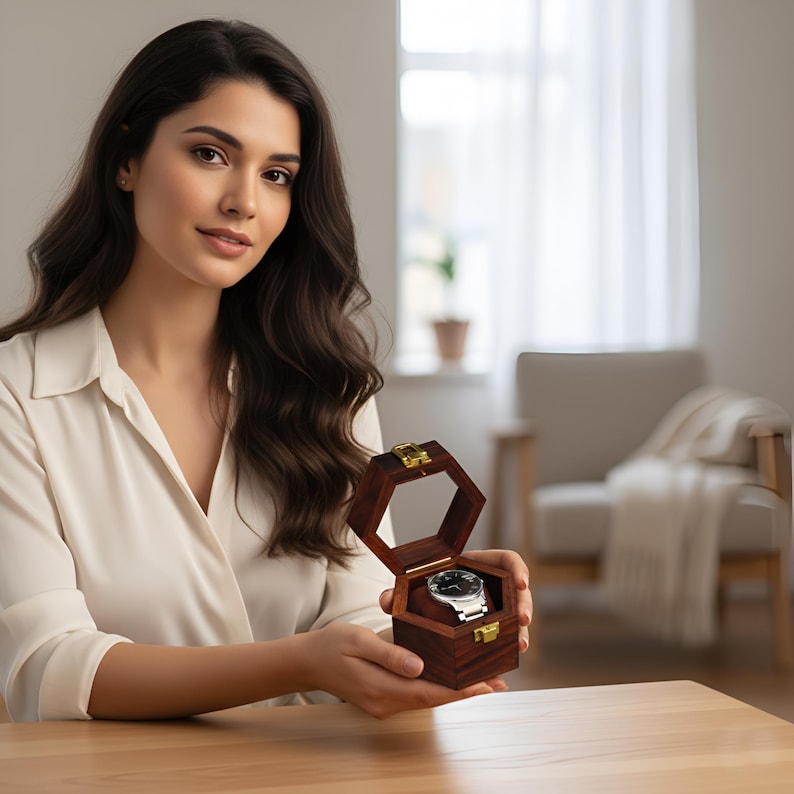 May include: A dark brown wooden hexagonal box with a hinged lid and gold-tone metal latch, containing a silver-tone wristwatch with a black face resting on a brown velvet cushion inside the open container.