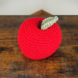 May include: A red crocheted apple with a green leaf on top. The apple is sitting on a wooden surface.