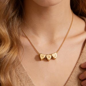 Könnte beinhalten: Eine goldfarbene Halskette mit drei herzförmigen Anhängern. Jeder Anhänger trägt einen einzelnen Buchstaben: H, R und J. Die Kette ist sichtbar, und die Halskette wird getragen. Das Design ist schlicht und elegant.