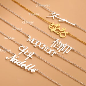 May include: A silver necklace with the name "Nadelle" written in cursive, along with the name written in different languages, including Korean, Russian, Chinese, Telugu, and Japanese. The necklace is on a light brown background.
