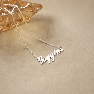 May include: A silver-colored necklace with the name "Goggins" in a cursive font. The necklace is on a light brown surface, with a decorative glass object in the background.