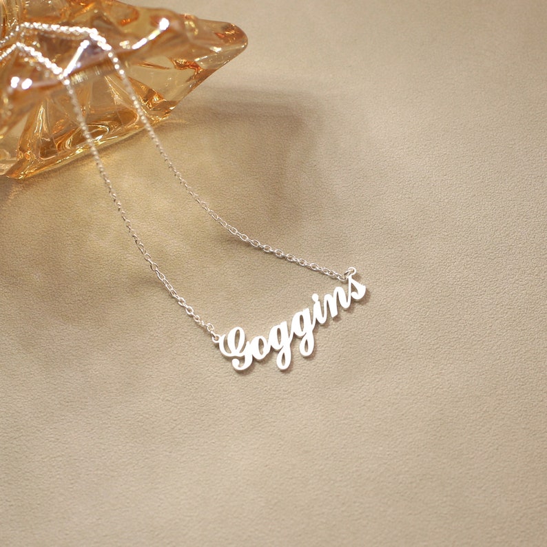 May include: A silver-colored necklace with the name "Goggins" in a cursive font. The necklace is on a light brown surface, with a decorative glass object in the background.
