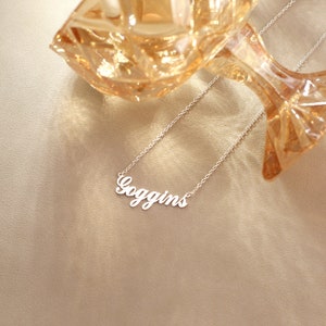 May include: A silver-colored necklace with the name "Goggins" in a cursive font. The necklace is displayed on a neutral-toned surface, with a decorative glass object in the background.