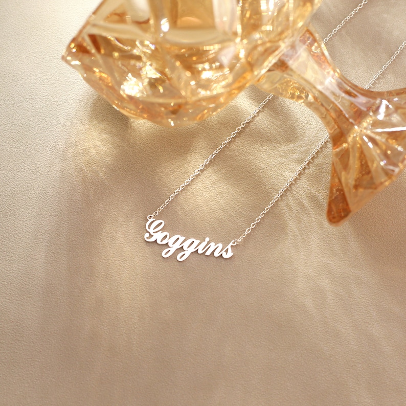 May include: A silver-colored necklace with the name "Goggins" in a cursive font. The necklace is displayed on a neutral-toned surface, with a decorative glass object in the background.