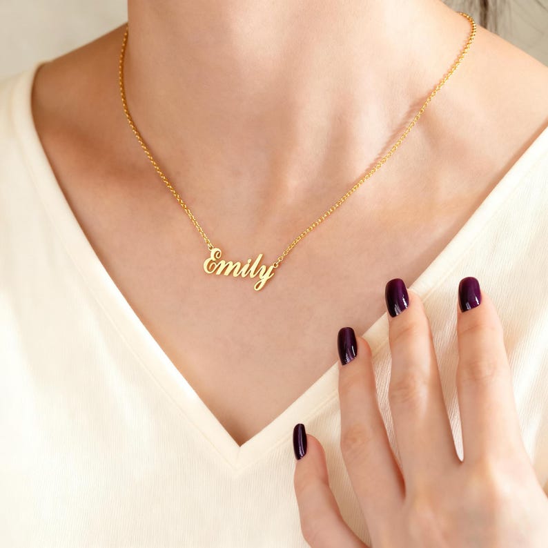 May include: A gold-colored necklace with the name "Emily" in cursive script. The necklace is worn around the neck. The person is wearing a cream-colored top. The person's nails are painted a dark purple color.