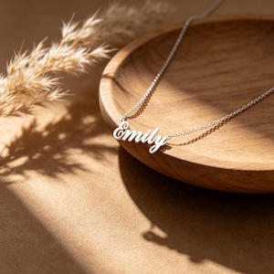 May include: A silver necklace with the name "Emily" in a cursive font. The necklace is displayed on a wooden bowl, with a neutral background. The chain is delicate and the name is the focal point.