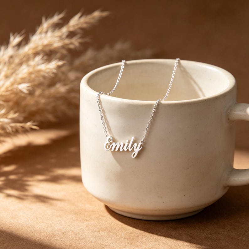 May include: A silver-colored necklace with the name "Emily" in cursive script. The necklace is resting inside a cream-colored mug. The background features dried plants and a brown surface.