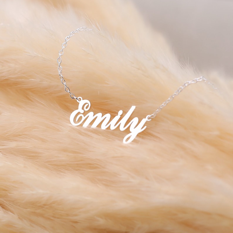 May include: A silver-colored necklace with the name "Emily" in a cursive font. The necklace is displayed against a soft, light brown background, creating a delicate and personalized jewelry piece.