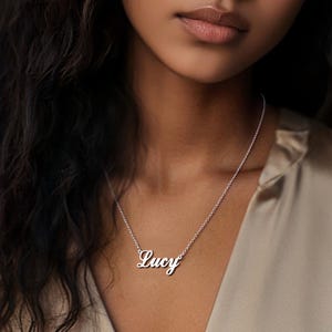 May include: A silver necklace with the name "Lucy" in a cursive font. The necklace is worn around the neck. The background is a soft, neutral color.