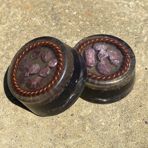 Könnte beinhalten: Zwei runde schwarze Untersetzer mit einem Kupferdraht-Rand. Jeder Untersetzer hat eine klare Harz-Oberfläche mit eingebetteten violetten Amethyst-Kristallen.