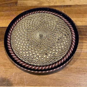 May include: A round coaster with a geometric pattern. The coaster has a dark rim, a copper-colored rope-like border, and a central design in shades of gold and brown. The coaster is on a wooden surface.