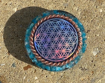 May include: A round, blue and copper-colored orgone energy device. The center features a holographic flower of life design. The device is surrounded by a copper coil and blue crystals, creating a unique, decorative piece.