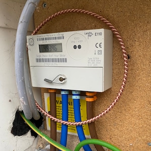 Könnte beinhalten: Ein weißer Landis+Gyr E110 Einphasen-Stromzähler mit einer Digitalanzeige, die 4939.6 anzeigt. Das Gerät verfügt über eine Kupferdrahtschleife, verschiedene elektrische Kabel in Blau, Braun und Gelb-Grün sowie ein gelbes Warnschild mit schwarzem Text.