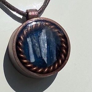 Puede incluir: Colgante redondo con marco de metal color cobre y centro de piedra azul. La piedra tiene un aspecto cristalino. El colgante cuelga de un cordón marrón.