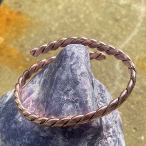 Puede incluir: Un brazalete de cobre con un diseño trenzado. El brazalete está hecho de una sola pieza de alambre de cobre que se ha trenzado para crear un efecto de cuerda.