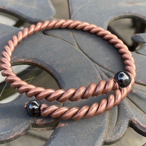 Puede incluir: Un brazalete de alambre de cobre con dos cuentas negras. El brazalete está trenzado y tiene un aspecto rústico.