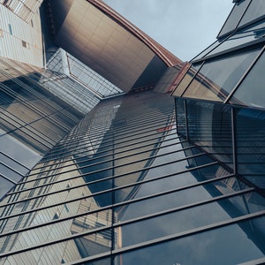 May include: Low-angle shot of a modern building with a complex geometric design. The building features reflective glass windows and a mix of gray and brown panels. The sky is overcast.