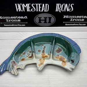 May include: A ceramic fish-shaped serving dish with a blue, green, and white color scheme. The dish has three compartments and is decorated with brown spots. The background includes a sign that says "Homestead Irons" and the Etsy shop URL.