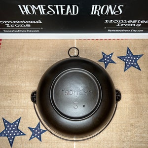 May include: A black cast iron Dutch oven with a handle. The bottom of the pot has the text "SIDNEY" and the number "3" embossed on it. The pot is sitting on a burlap fabric with red and white stripes.