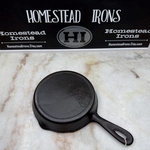 May include: A black cast iron skillet with a single handle and a loop for hanging. The skillet is round with a slightly raised edge. The background includes a sign that says "Homestead Irons".