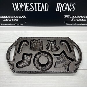 May include: A black cast iron Christmas cookie mold with seven different designs: a candy cane, a present, a bell, a wreath, a stocking, a star, and a Christmas tree. The mold is made by Homestead Irons.