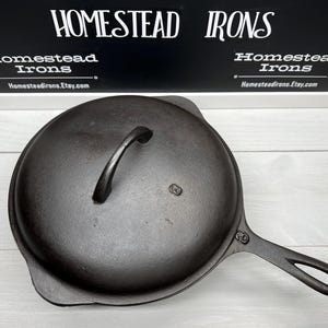 May include: A black cast iron skillet with a lid and a handle. The skillet has a smooth surface and is marked with the number "6" on the lid.