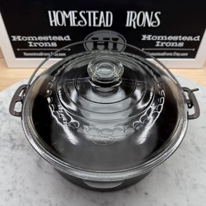 May include: A clear glass casserole dish with a matching lid. The dish has a dark gray base and handles. The lid has a clear glass knob. The text "HOMESTEAD IRONS" is visible in the background.