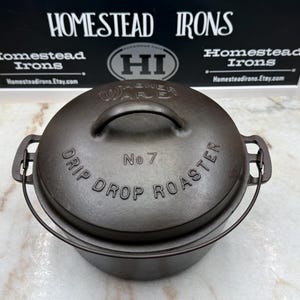May include: A dark brown cast iron drip drop roaster with a lid and handle. The lid is inscribed with "Drip Drop Roaster" and "No 7". The product is from Homestead Irons, as indicated by the sign in the background.
