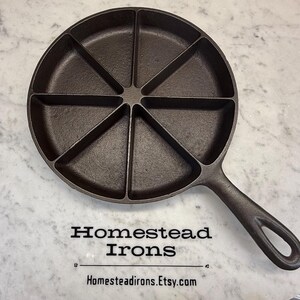 May include: A dark brown cast iron cornbread pan with a handle and seven triangular sections. The pan is on a white marble surface. The text "Homestead Irons" and "Homesteadirons.Etsy.com" are printed below the pan.